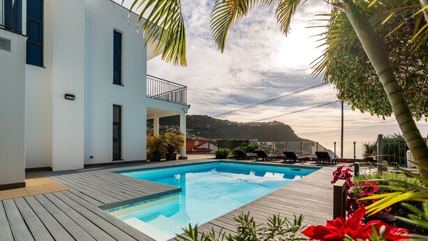 A swimming pool with a wooden deck, surrounded by plants and a white building with a balcony.