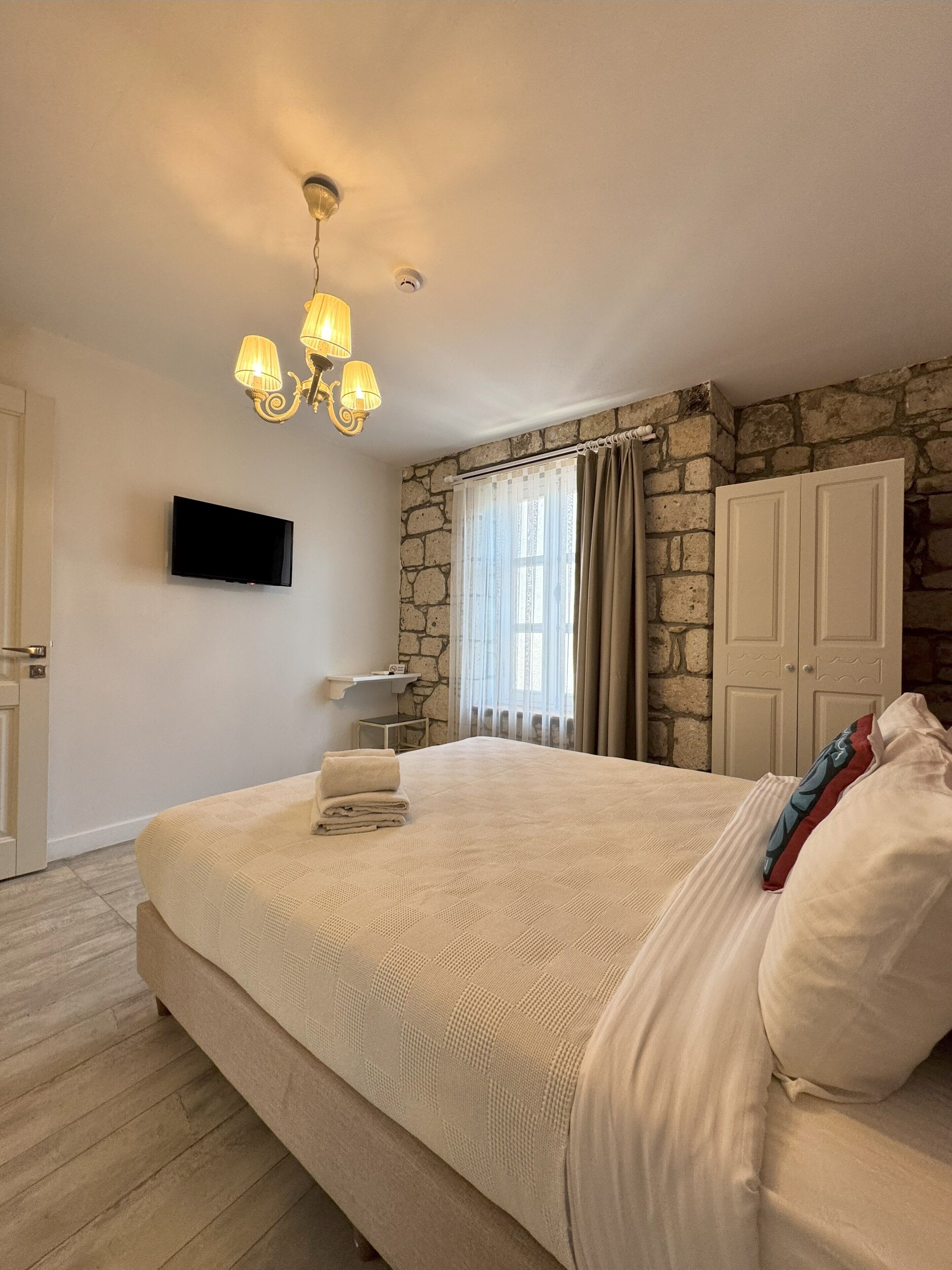 A bedroom with a large bed, a flat-screen TV mounted on the wall, a stone accent wall, a chandelier, and a window with curtains.
