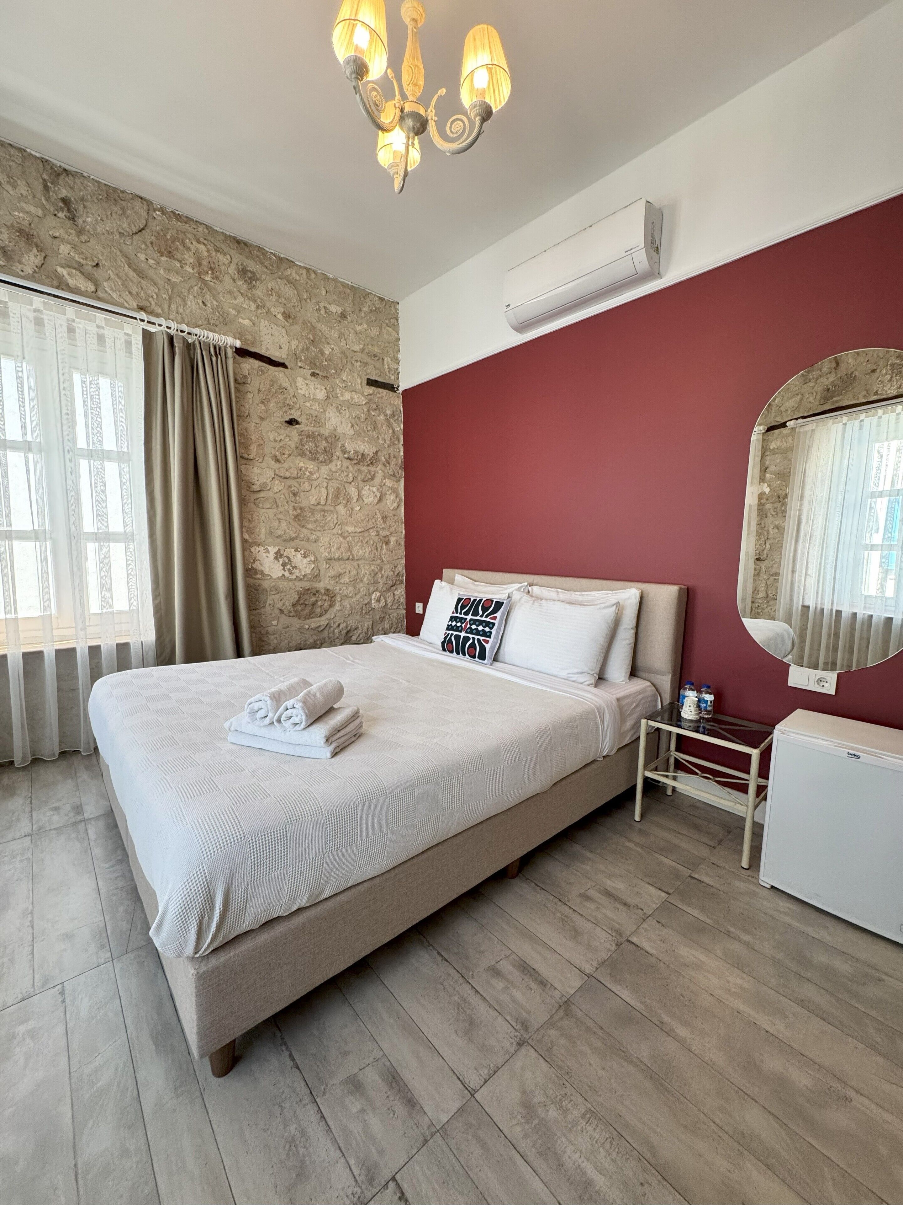 Een hotelkamer met een groot bed, een stenen muur, een rode accentmuur, een kroonluchter en een kleine tafel met een spiegel.