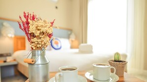 Quarto de hotel com cama, mesa de cabeceira, mesinha de centro com duas xícaras de café, vaso de metal com flores secas e um pequeno cacto em vaso.