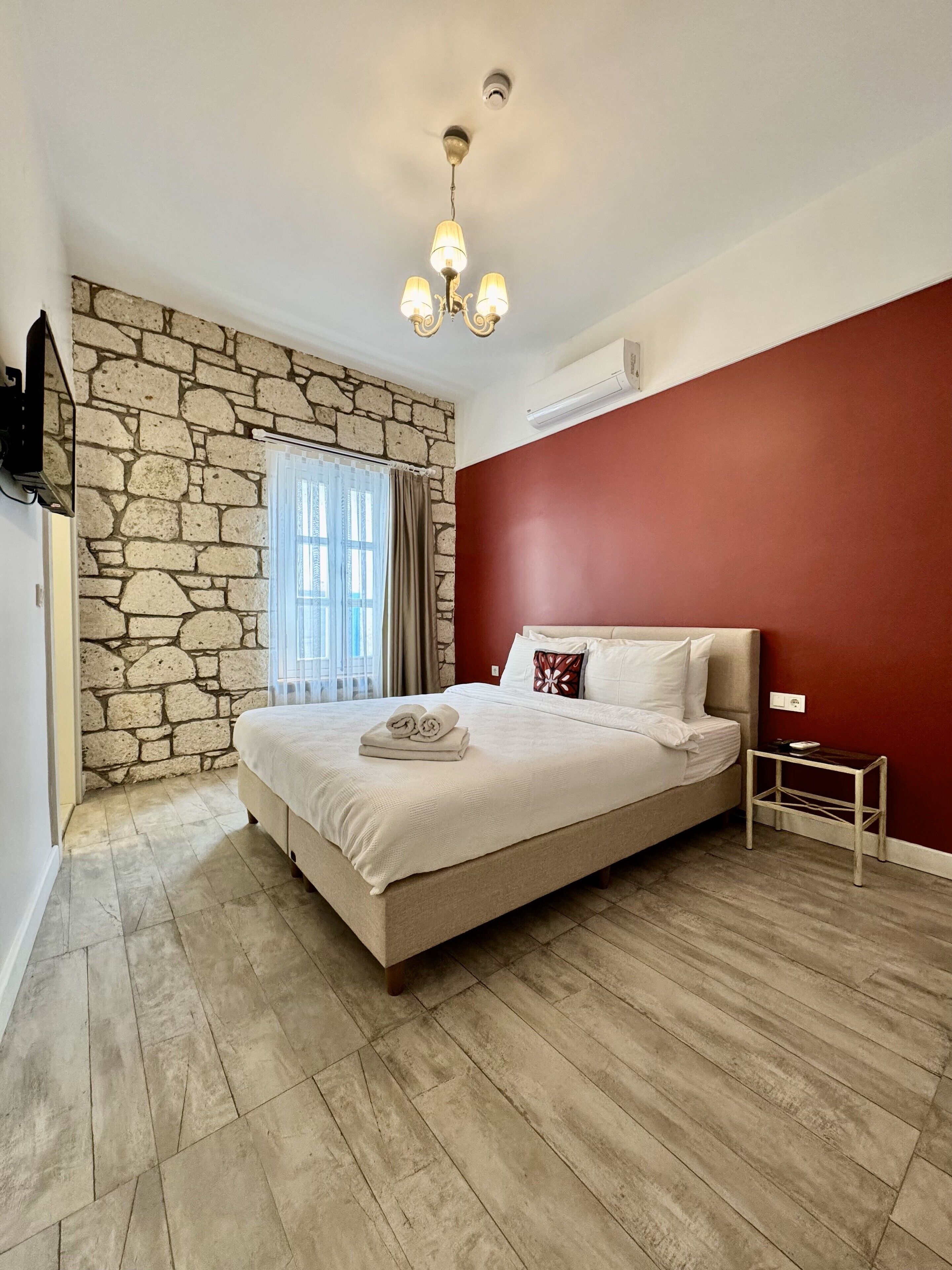 A bedroom with a stone wall, a bed with white linens, a red accent wall, and a ceiling light fixture.
