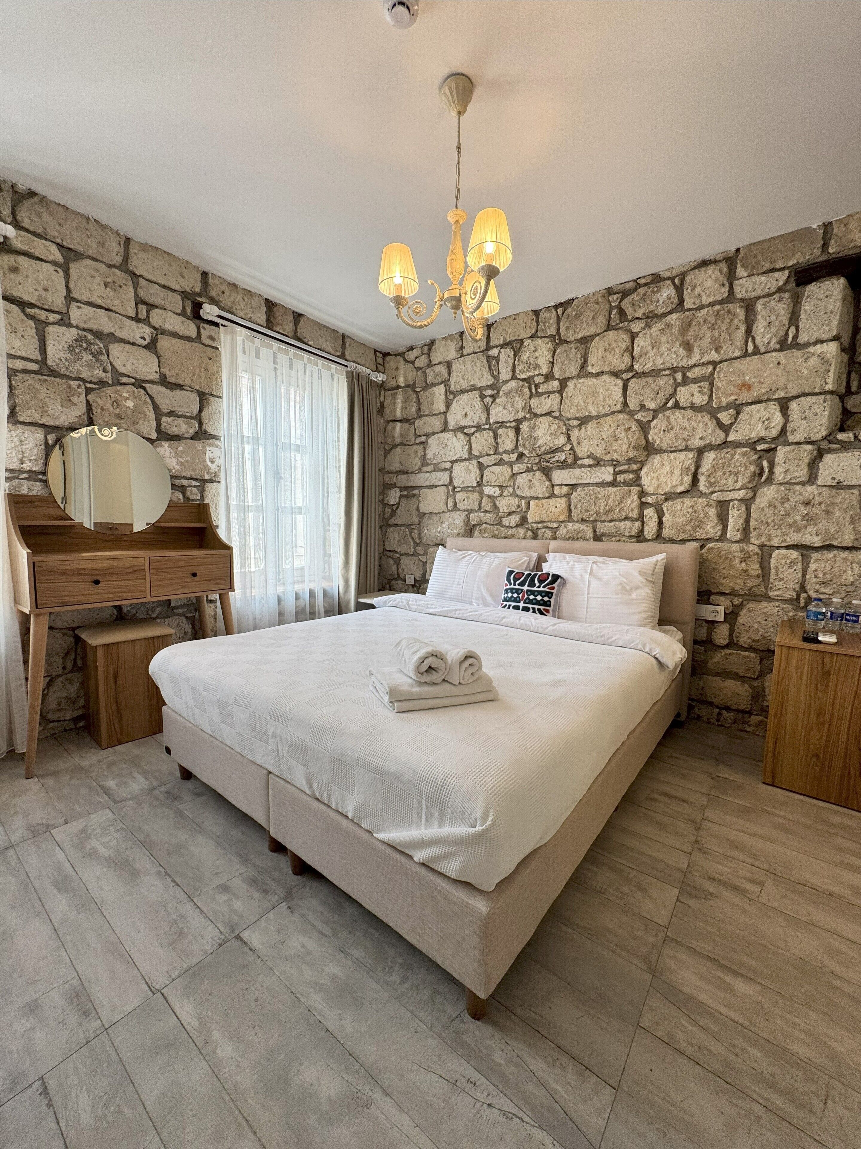 Un dormitorio con pared de piedra, una cama con sábanas blancas, un escritorio de madera, un espejo y una araña.