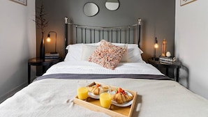 Un lit bien fait, agrémenté d’un coussin à motifs floraux, une table avec un plateau de petit-déjeuner comprenant des croissants et du jus d’orange, et des lampes de chevet.