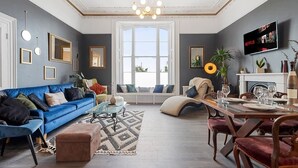 Un salon spacieux avec un canapé bleu, une table à manger dressée pour six personnes et une grande fenêtre laissant entrer la lumière naturelle.