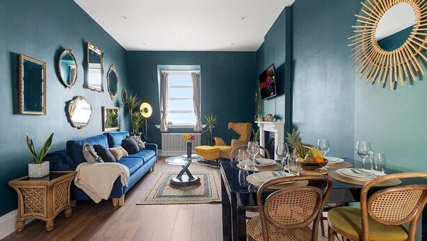Una sala de estar con un sofá azul, una mesa de comedor y sillas, un televisor y un espejo decorativo.