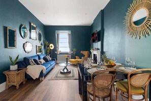 Una sala de estar con un sofá azul, una mesa de comedor y sillas, un televisor y un espejo decorativo.