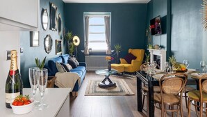 Una sala de estar moderna con una pared azul, un sillón amarillo, una mesa de comedor preparada para cuatro comensales y un ventanal con cortinas.