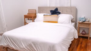 A neatly made bed with white bedding, a wooden nightstand with a lamp, and a small table with a vase and a clock.