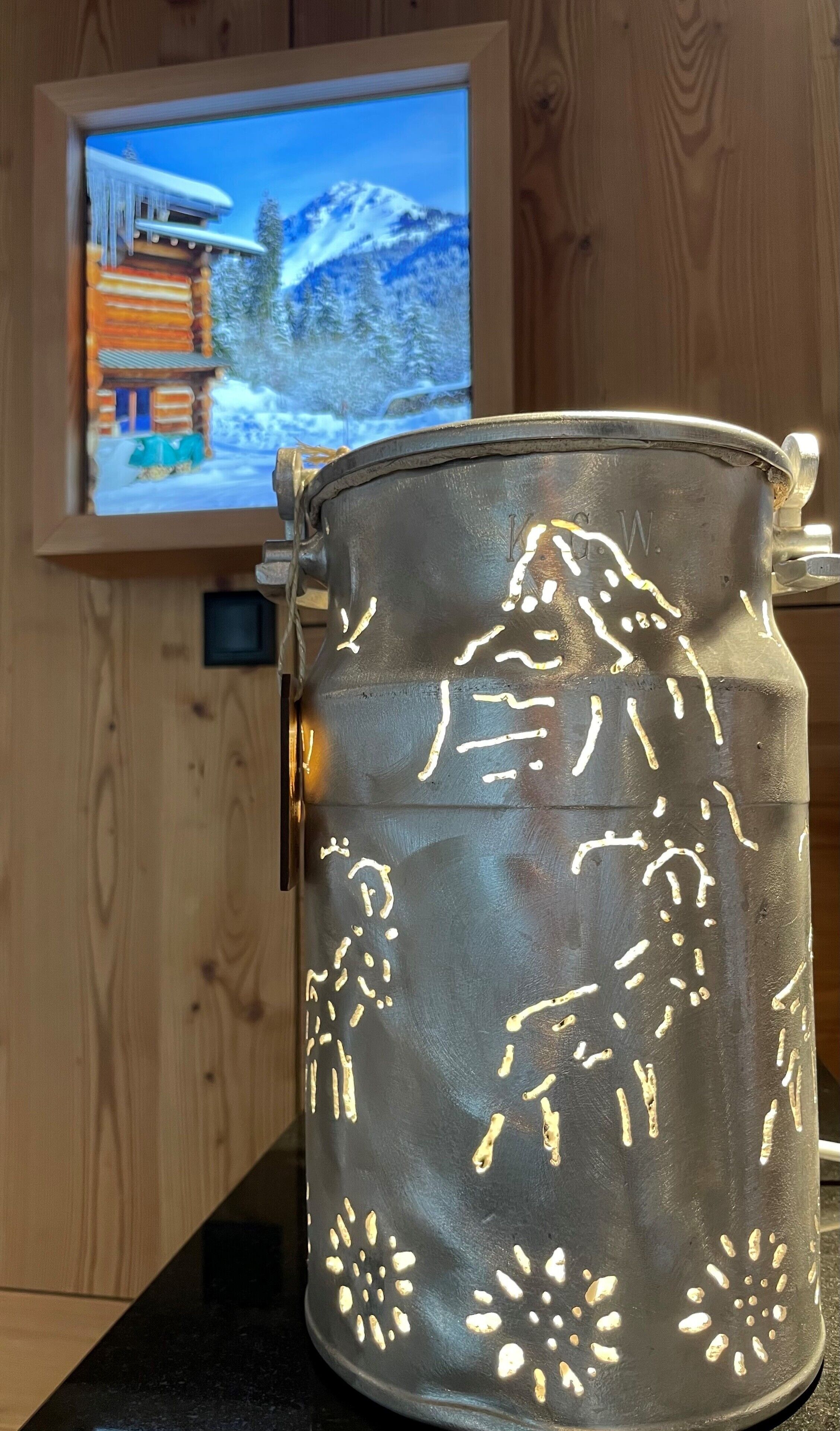 A metal container with embossed designs, placed on a wooden surface with a television displaying a snowy landscape in the background.