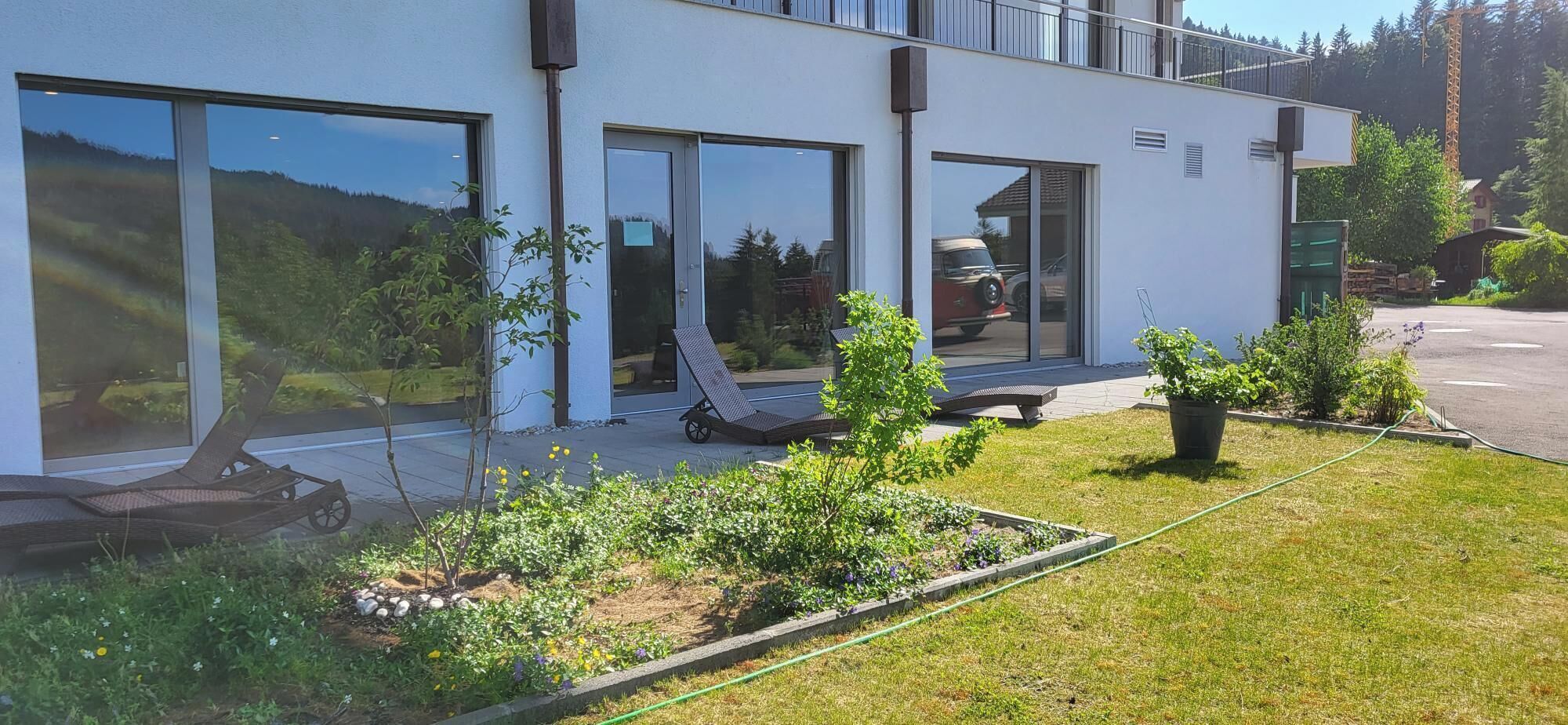 Un bâtiment moderne doté de grandes fenêtres, d’un espace jardin avec des plantes et une pelouse, ainsi que de sièges extérieurs.