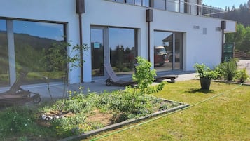 Un bâtiment moderne doté de grandes fenêtres, d’un espace jardin avec des plantes et une pelouse, ainsi que de sièges extérieurs.