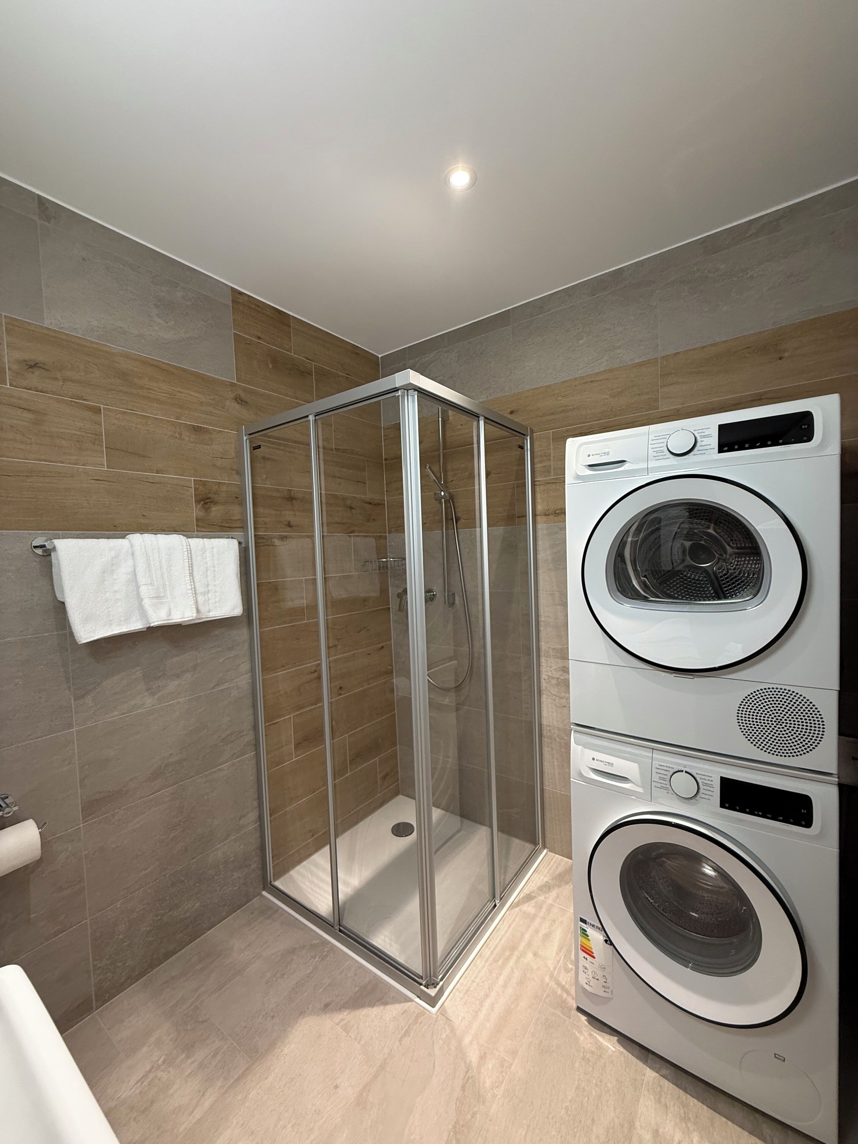 Une salle de bain moderne dotée d’une douche à parois de verre, d’un lave-linge et d’un sèche-linge superposés, et d’un porte-serviettes sur lequel est posée une serviette blanche.