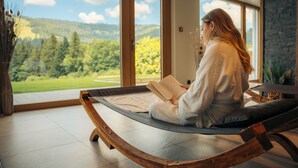 A person sitting on a wooden swing chair reading a book in a room with large windows overlooking a green landscape. - Boutique Hôtel Corbetta (Chatel-Saint-Denis)