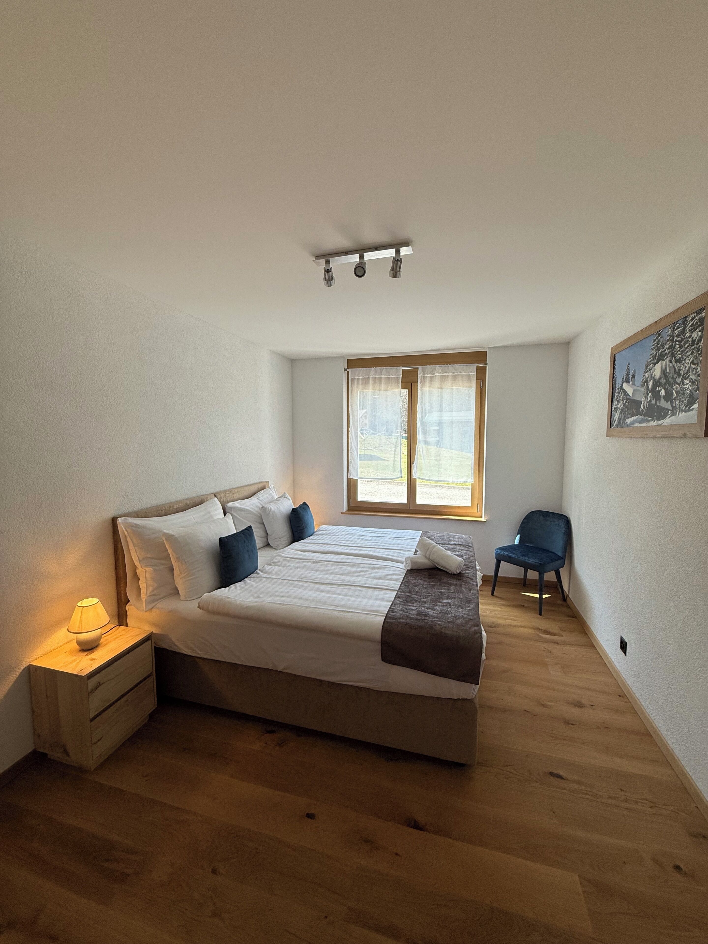 Un dormitorio con una cama grande, una mesita de noche con lámpara, una ventana con cortinas y una silla.