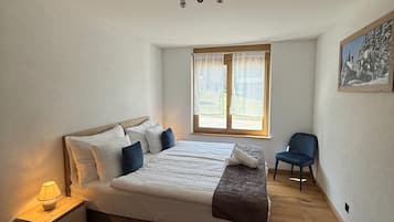 A bedroom with a large bed, a bedside table with a lamp, a window with curtains, and a chair.