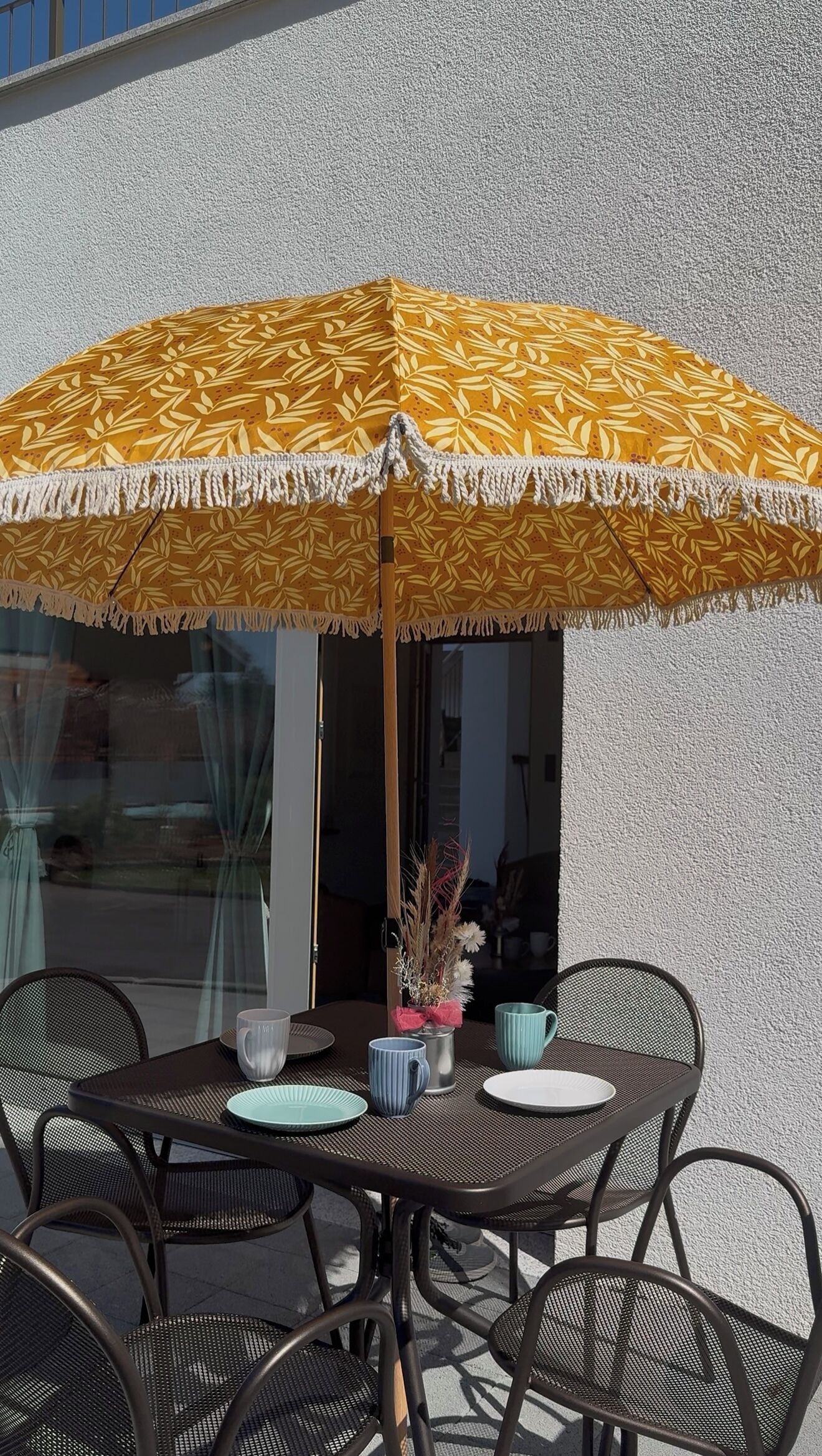 Et udendørs spiseomr åde med en gul mønstret parasol, metalstole og et bord dækket med tallerkener, kopper og en vase med blomster.