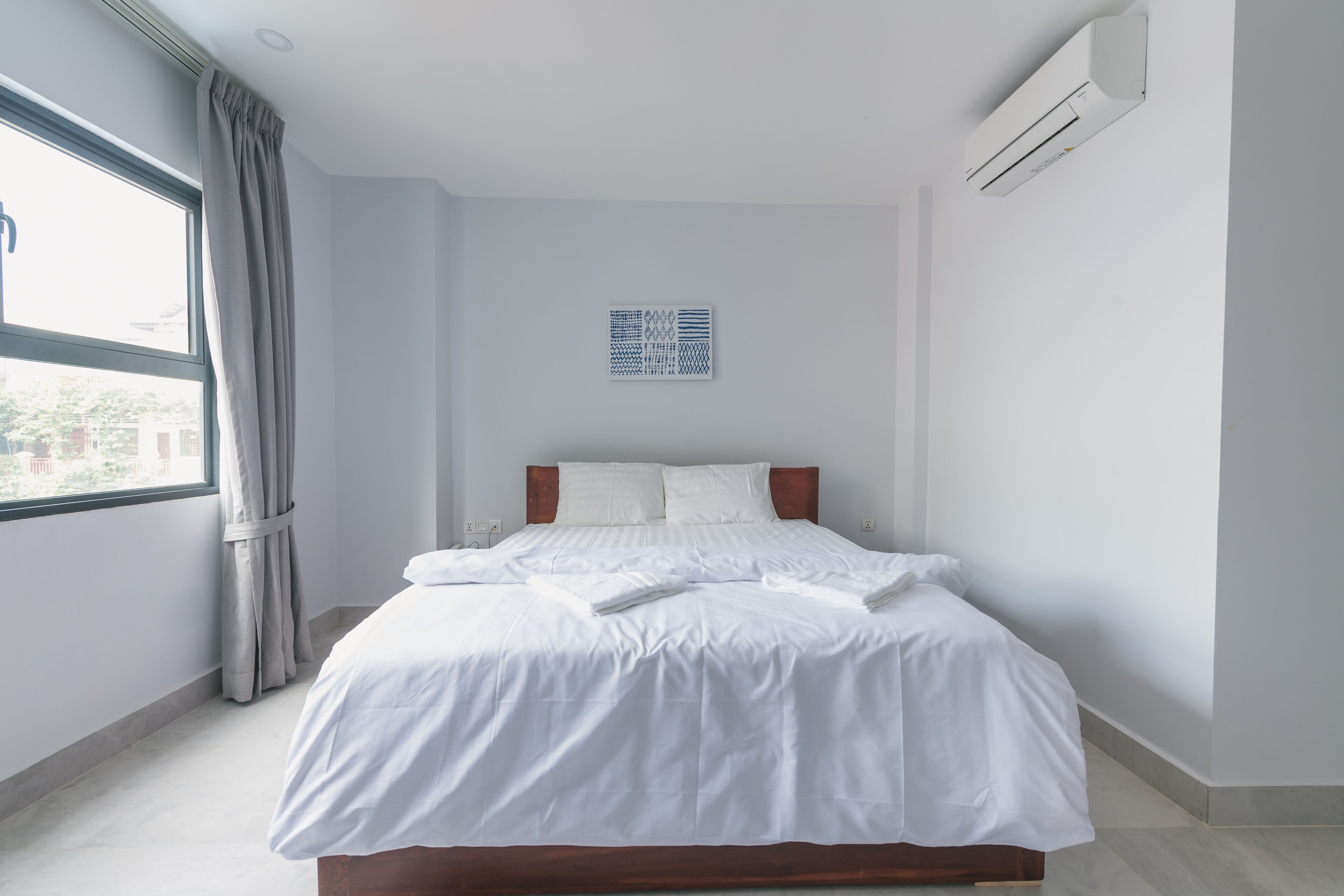 A modern hotel room with a large bed, a window with curtains, and a wall-mounted air conditioner.