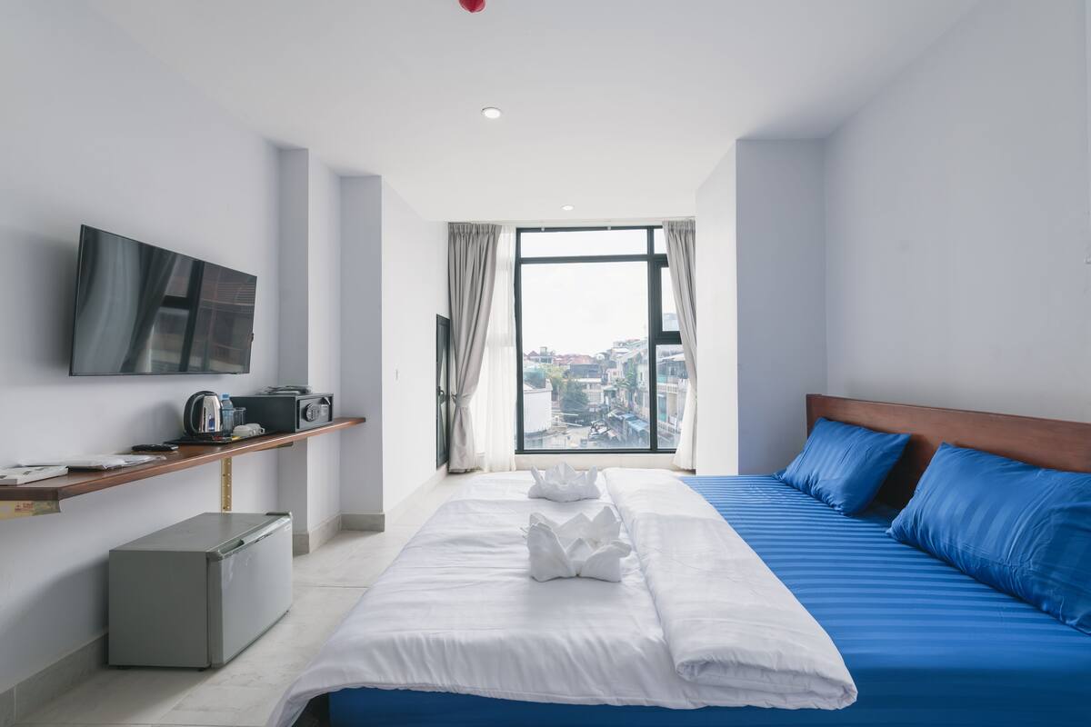 A modern hotel room with a large bed, a TV mounted on the wall, a small kitchenette, and a city view through the window.