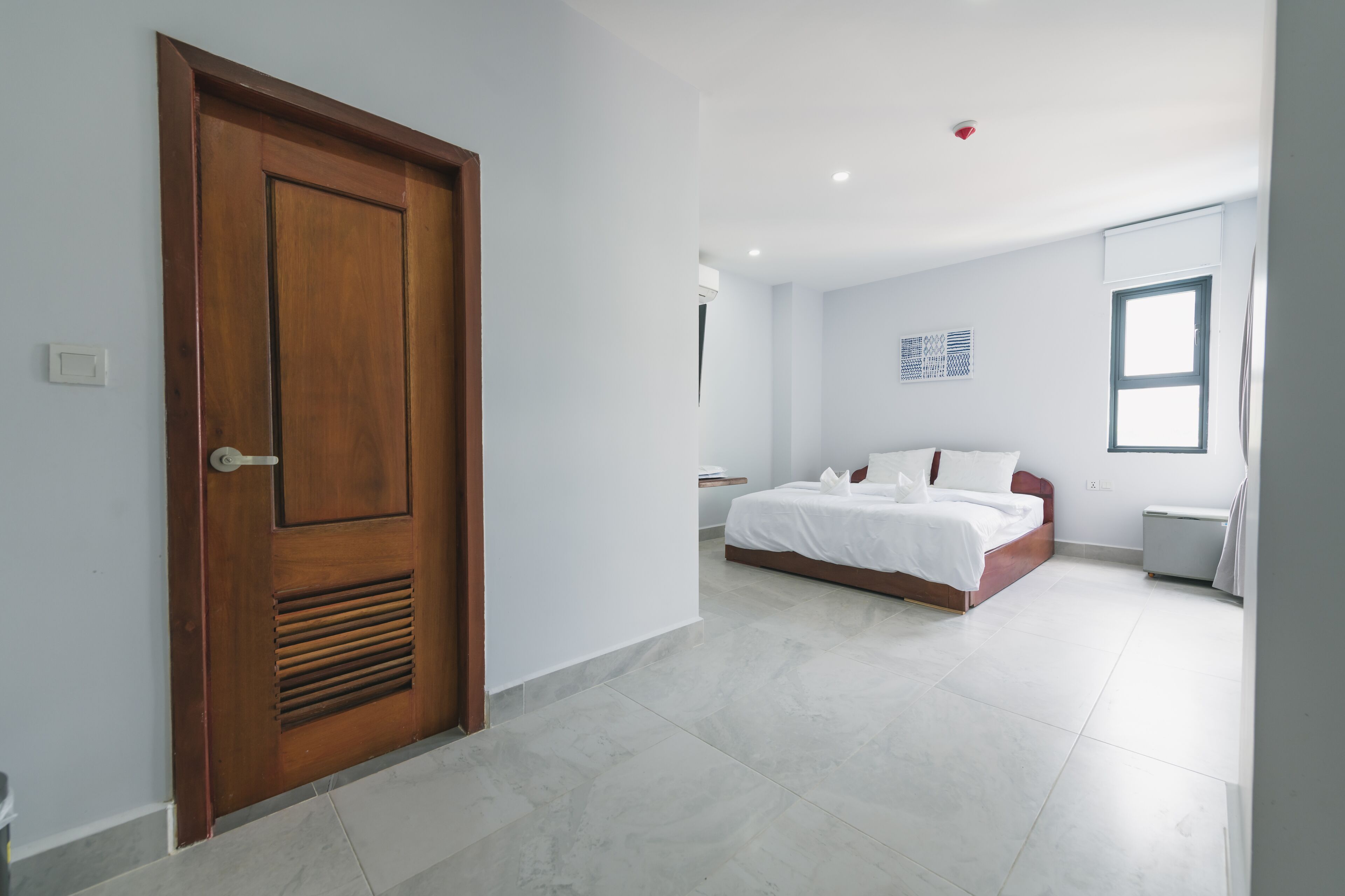 A bedroom with a wooden door, a bed, a bedside table, a window, and a ceiling light.