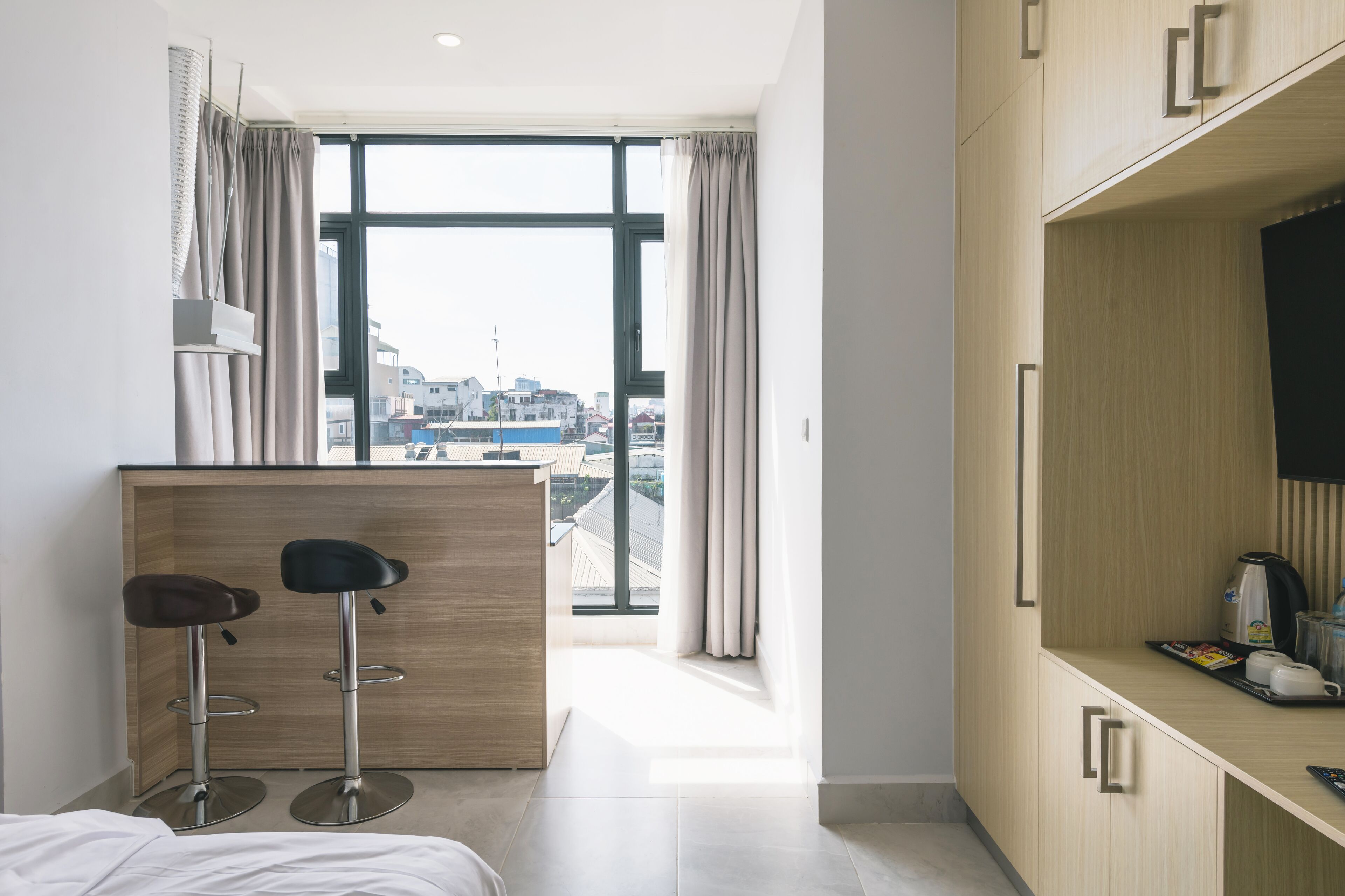 A modern hotel room with a large window, a wooden desk with two bar stools, a bed, and a kitchenette area.