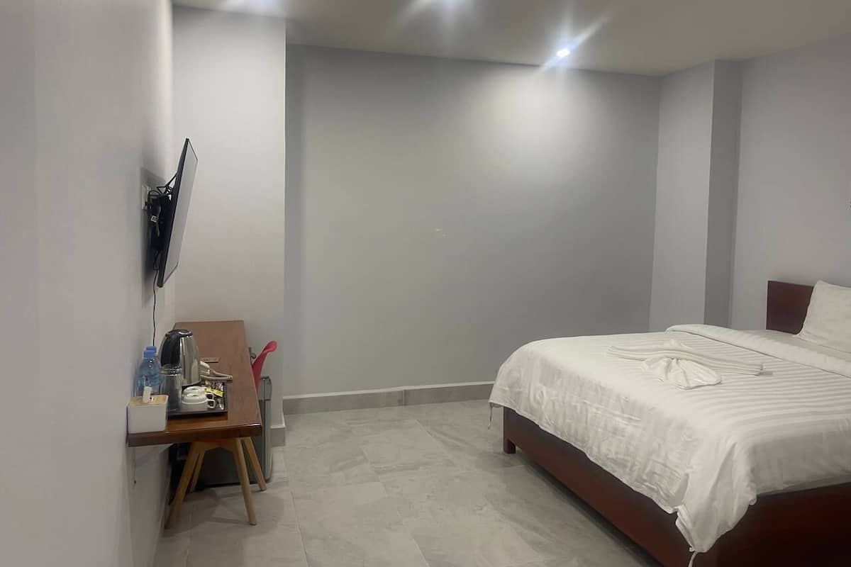 A bedroom with a bed, a desk with a chair, a flat-screen TV mounted on the wall, and a small table with a kettle and water bottle.