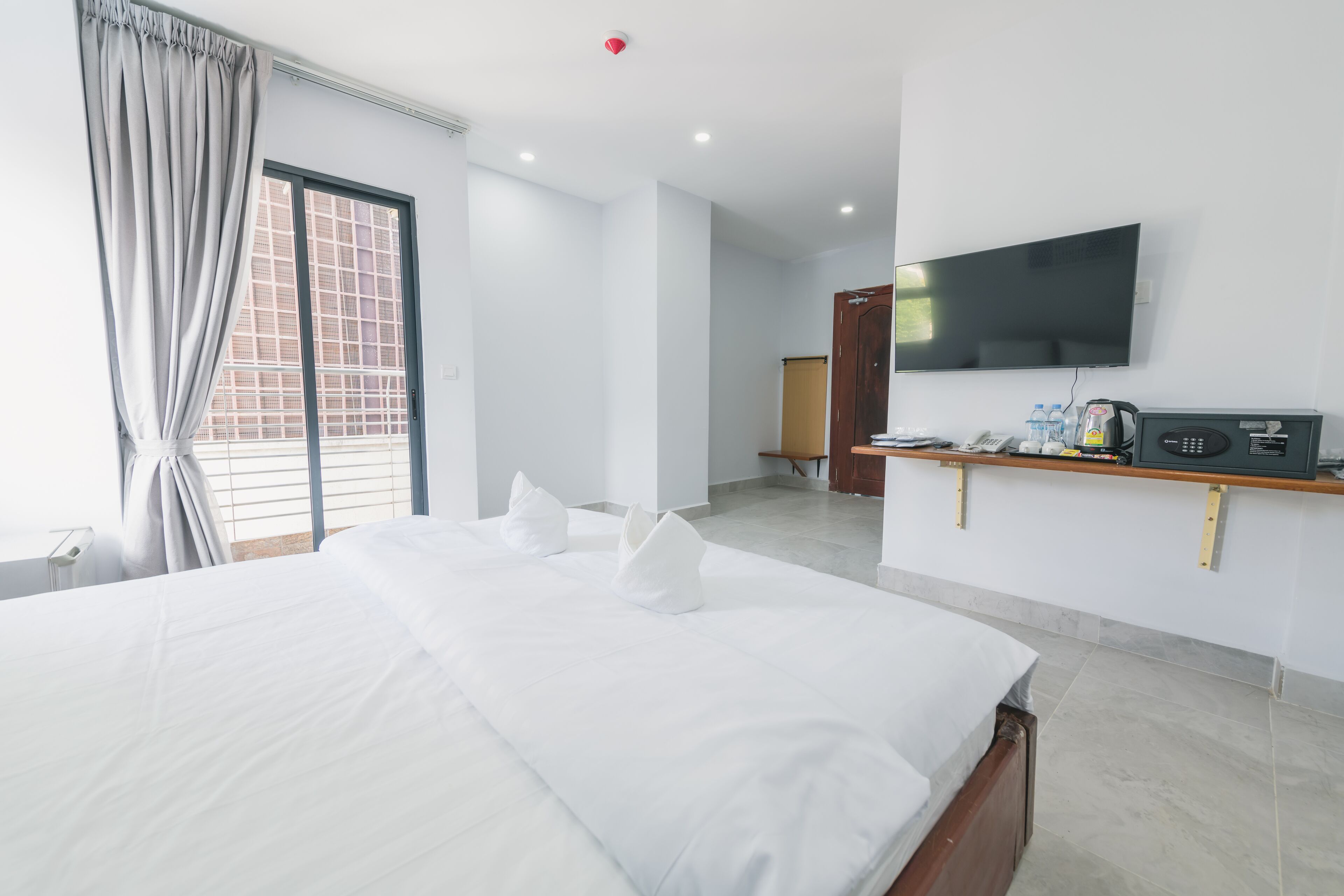 A hotel room with a large bed, a flat-screen TV mounted on the wall, a wooden side table with a coffee maker, and a window with curtains.