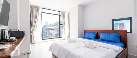 Habitación de hotel con una cama grande, un escritorio con hervidor y vistas de la ciudad a través de un ventanal amplio.