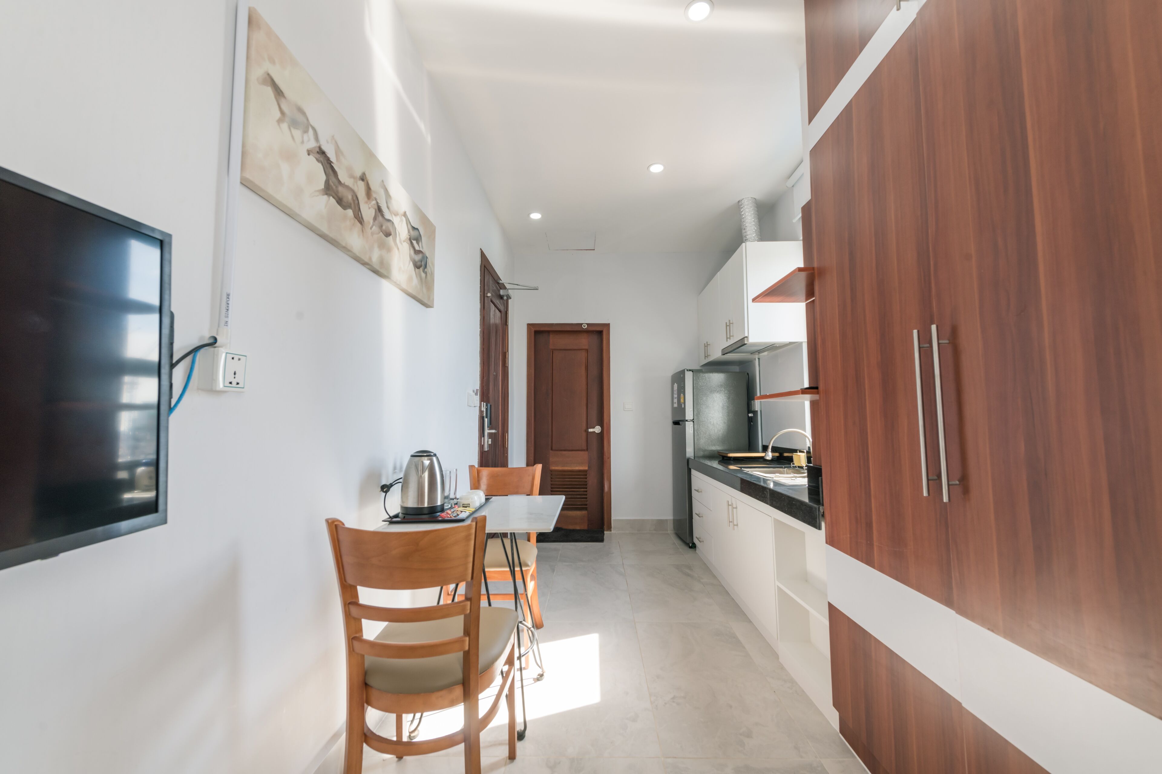 A narrow kitchen with wooden cabinets, a dining table with chairs, and a television mounted on the wall.