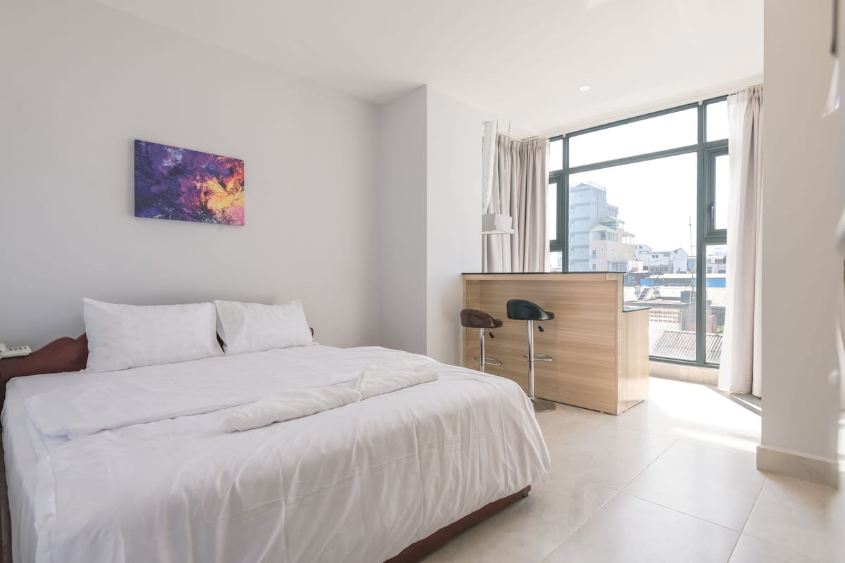 A modern hotel room with a large bed, a desk with two chairs, a painting on the wall, and a view of the city through large windows.