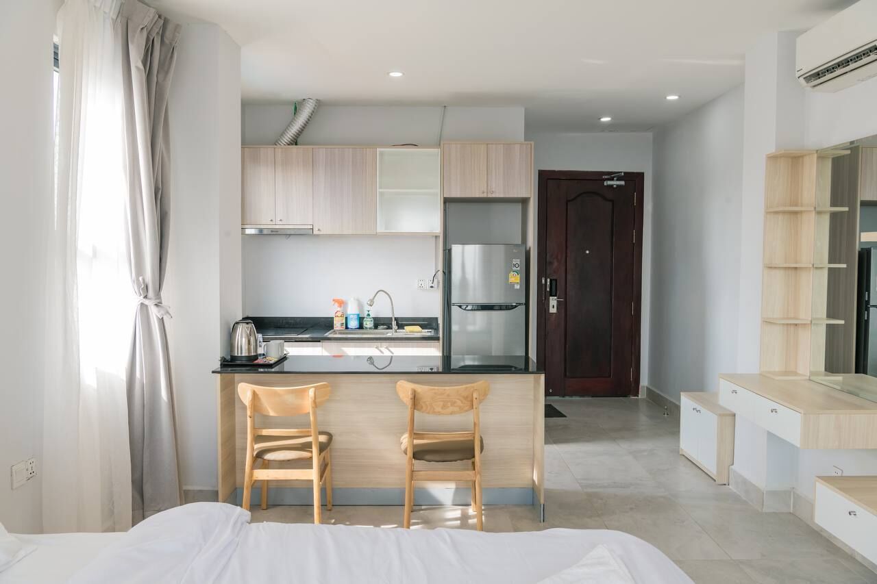 A modern hotel room with a kitchenette, two wooden chairs, a bed, and a dark wooden door.
