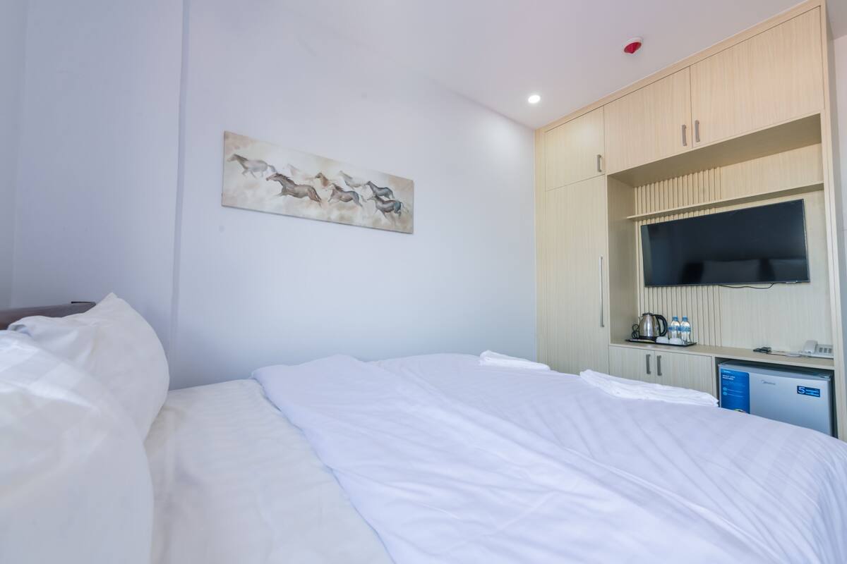 A hotel room with a bed, a painting on the wall, a TV, and a minibar.