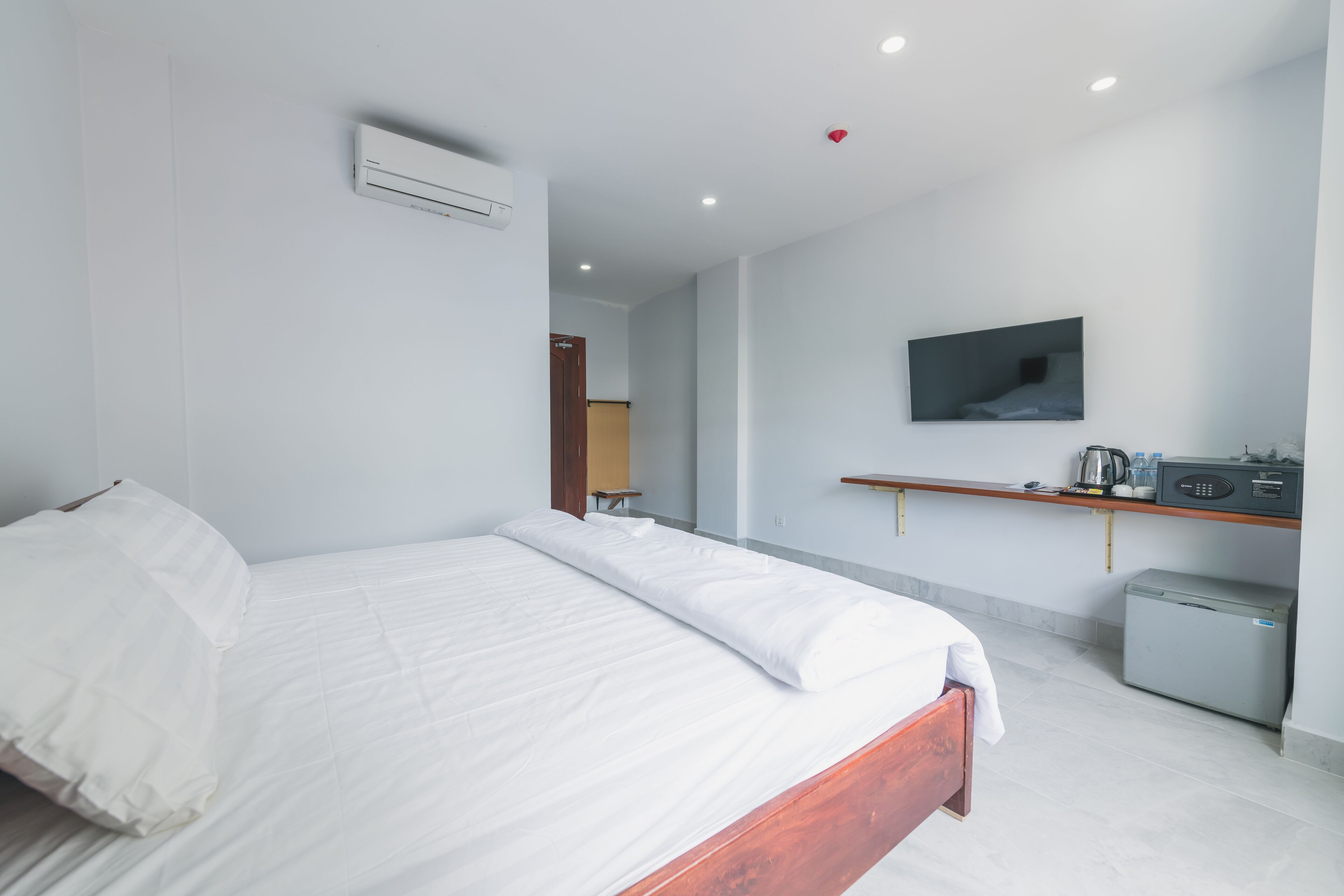 A modern hotel room with a large bed, a television mounted on the wall, a small refrigerator, and a wooden shelf with a kettle and other items.