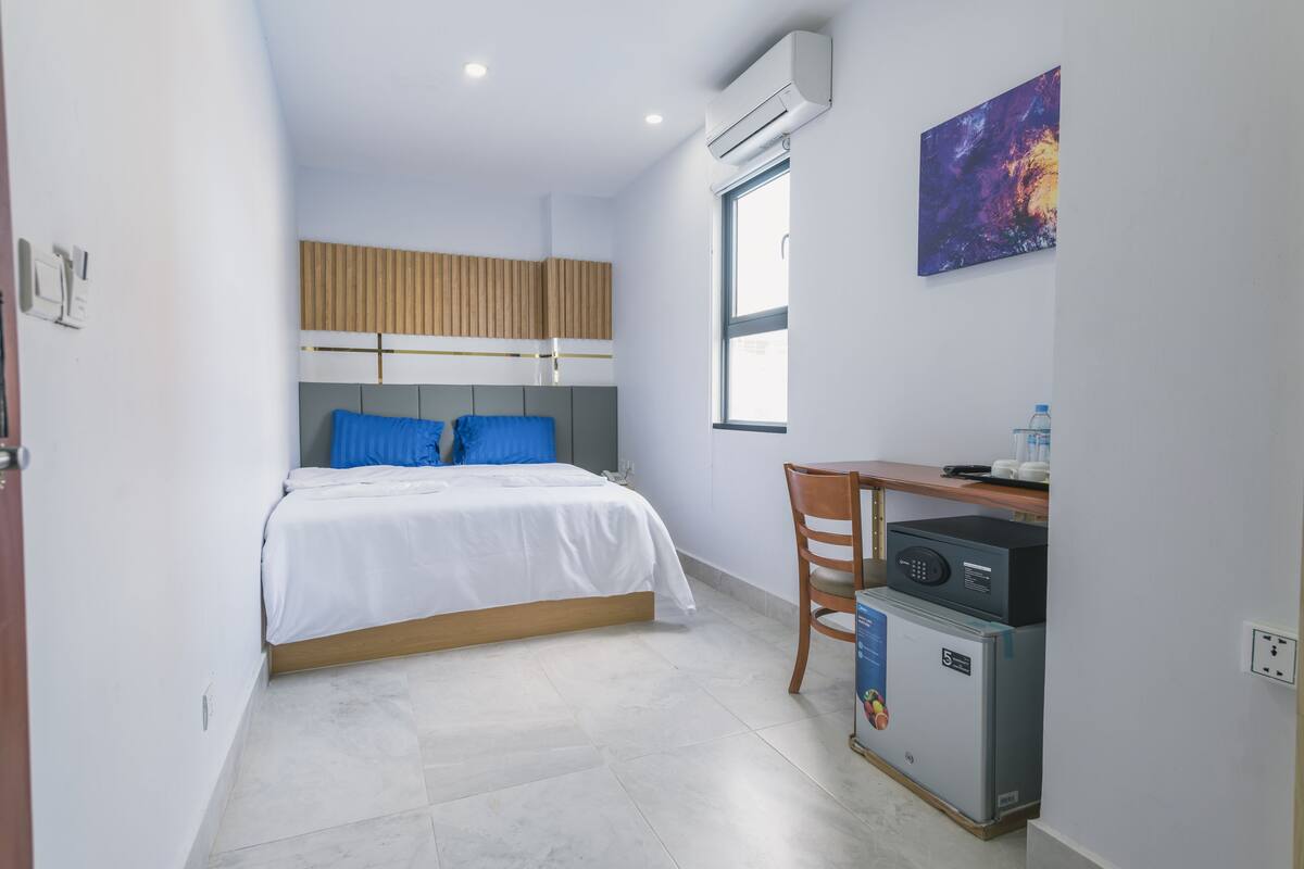 A compact hotel room with a bed, a desk, a chair, a small refrigerator, and a window.