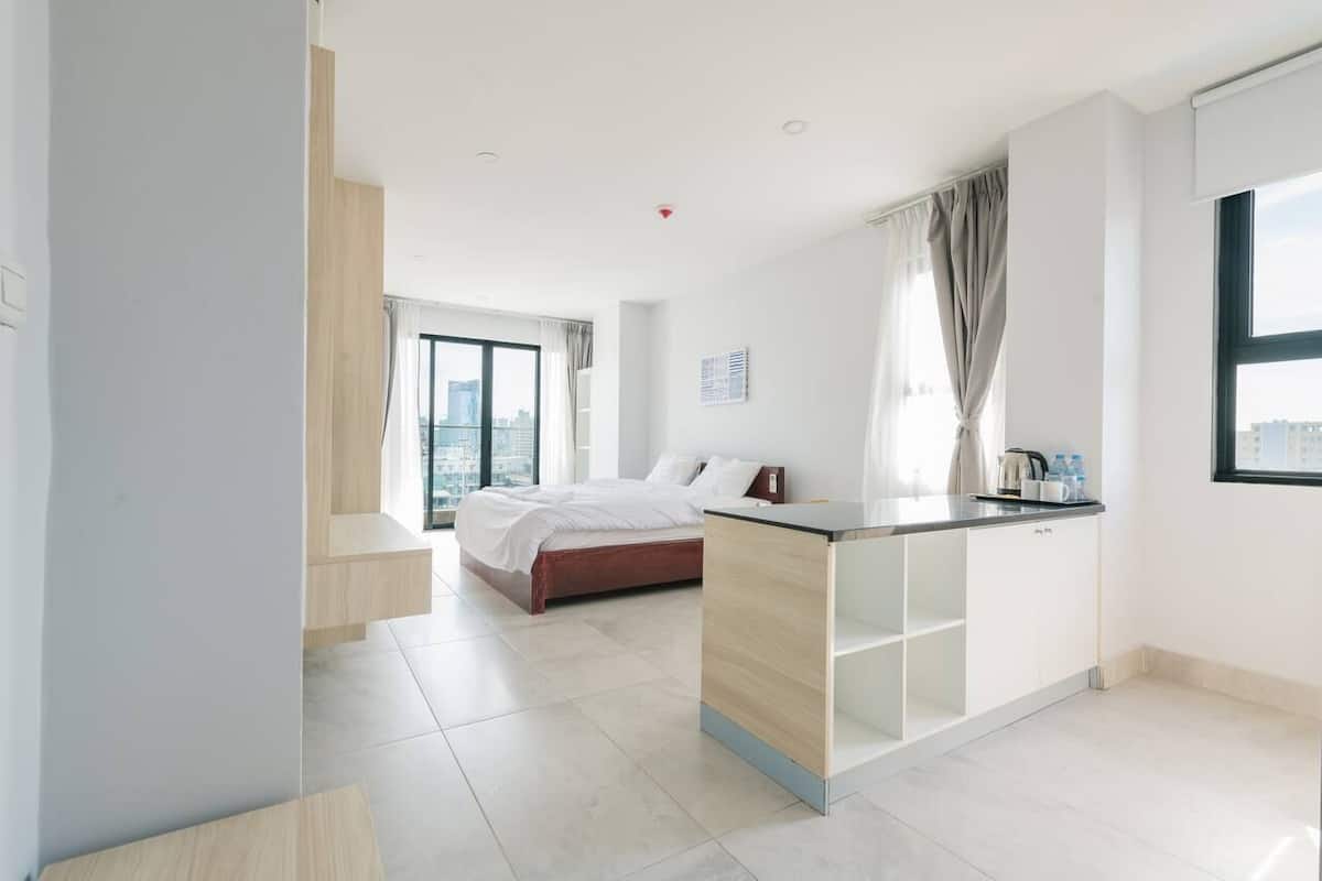 A modern hotel room with a bed, a kitchenette, a large window, and a city view.