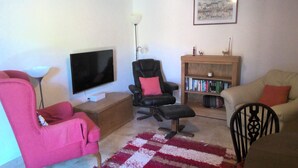 Un salon comprenant un téléviseur à écran plat, un fauteuil de bureau noir, un fauteuil rouge, un canapé beige et une bibliothèque remplie de livres.
