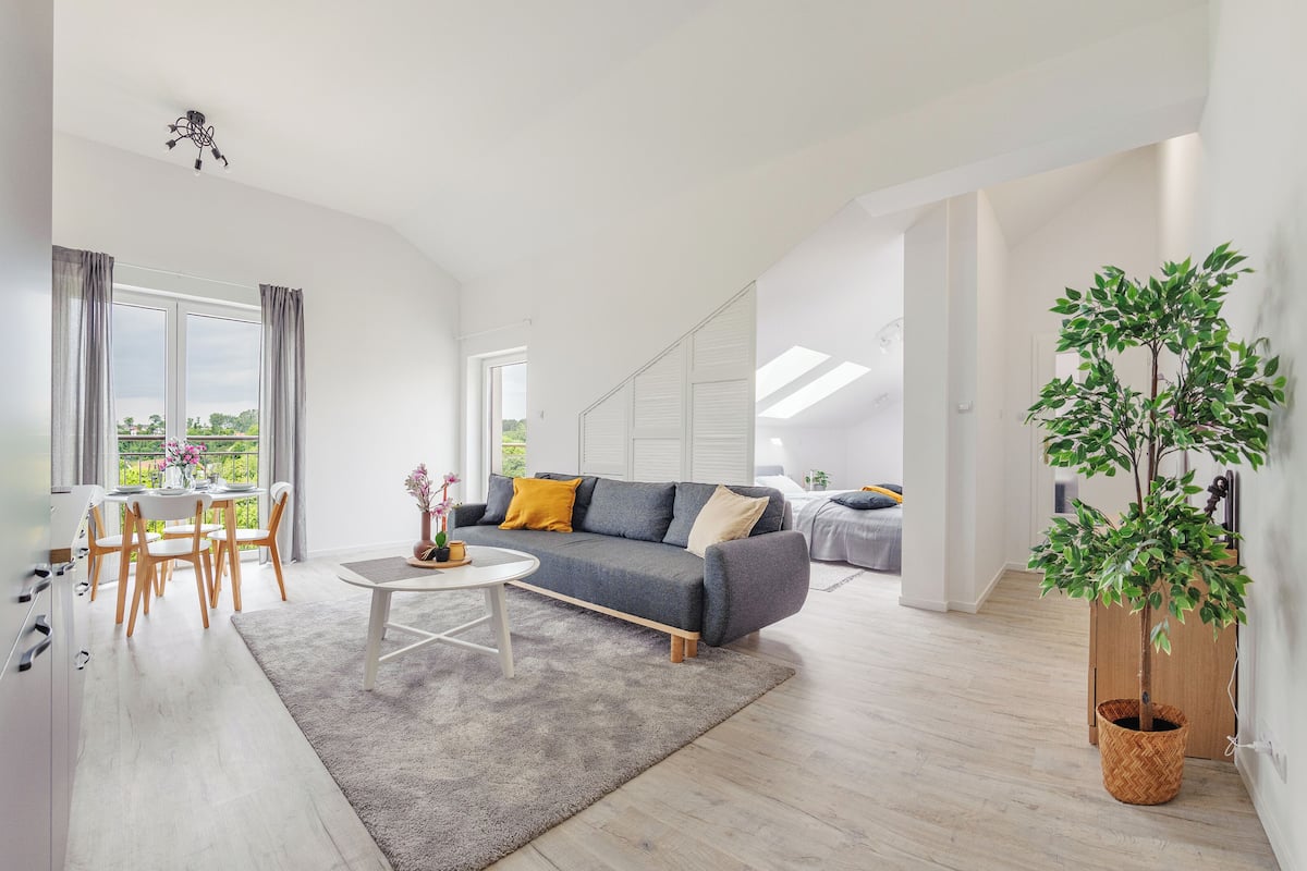 A modern living room with a grey sofa, a dining table with chairs, and a large window with curtains.