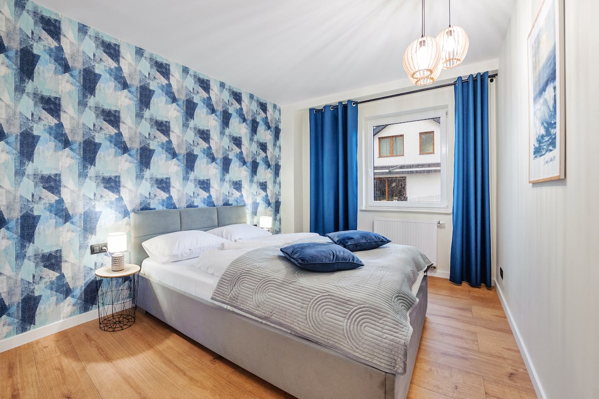 A bedroom with a bed, a bedside table with a lamp, a window with curtains, and a wall with a geometric pattern.