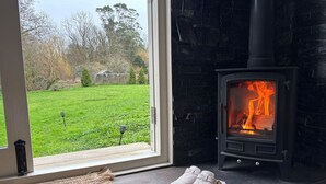 A cozy room with a stone fireplace, a pair of slippers on the floor, and a view of a green garden through a glass door.