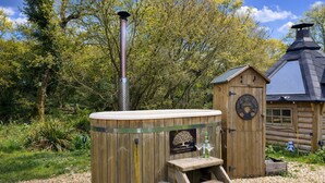 A wooden outdoor sauna with a metal chimney, a wooden bench, and a small table with a bottle and glass.