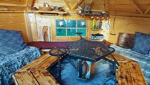 A cozy cabin interior with a central wood-burning stove, a window with a view of greenery, and a wooden ceiling.