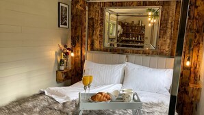 A bed with a tray of croissants, juice, and coffee cups, a fluffy rug, a wooden wall with a mirror, and a small shelf with a bottle and flowers.
