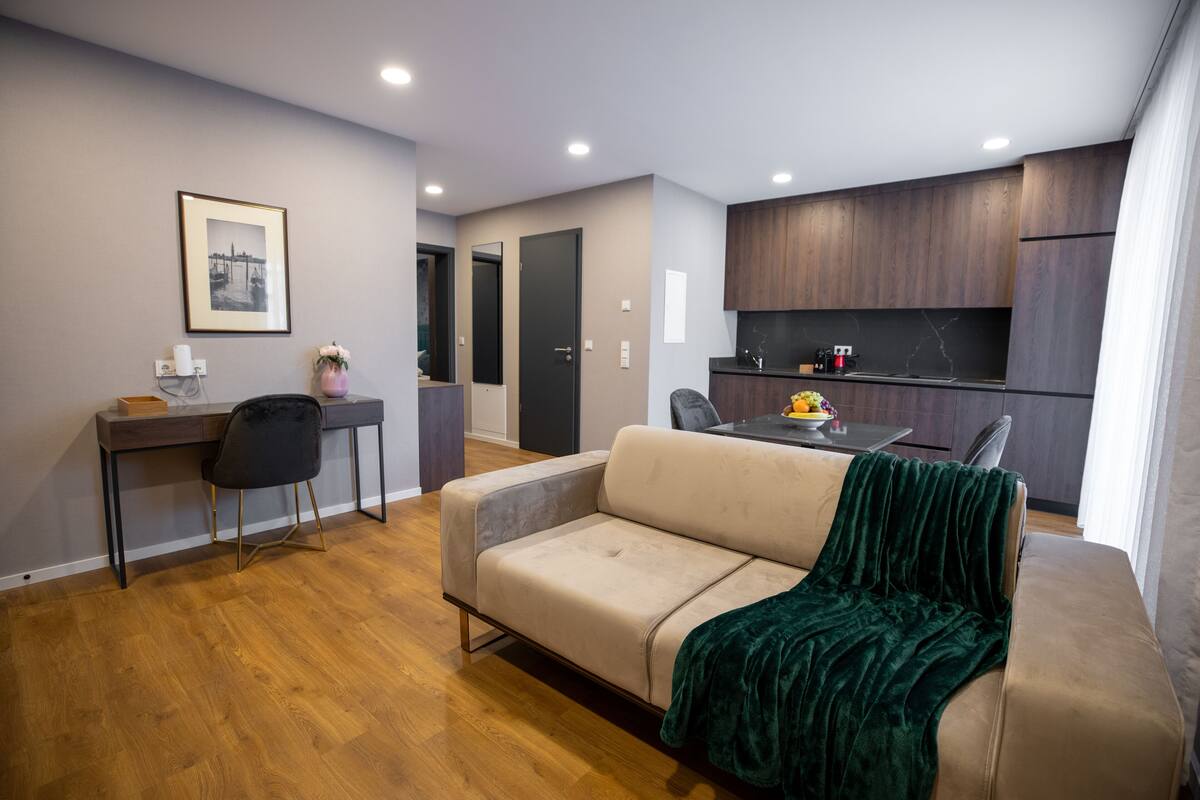 Una sala moderna con un sofá, un escritorio y una silla. Hay una zona de cocina con armarios de madera oscura y una mesa de comedor con sillas.