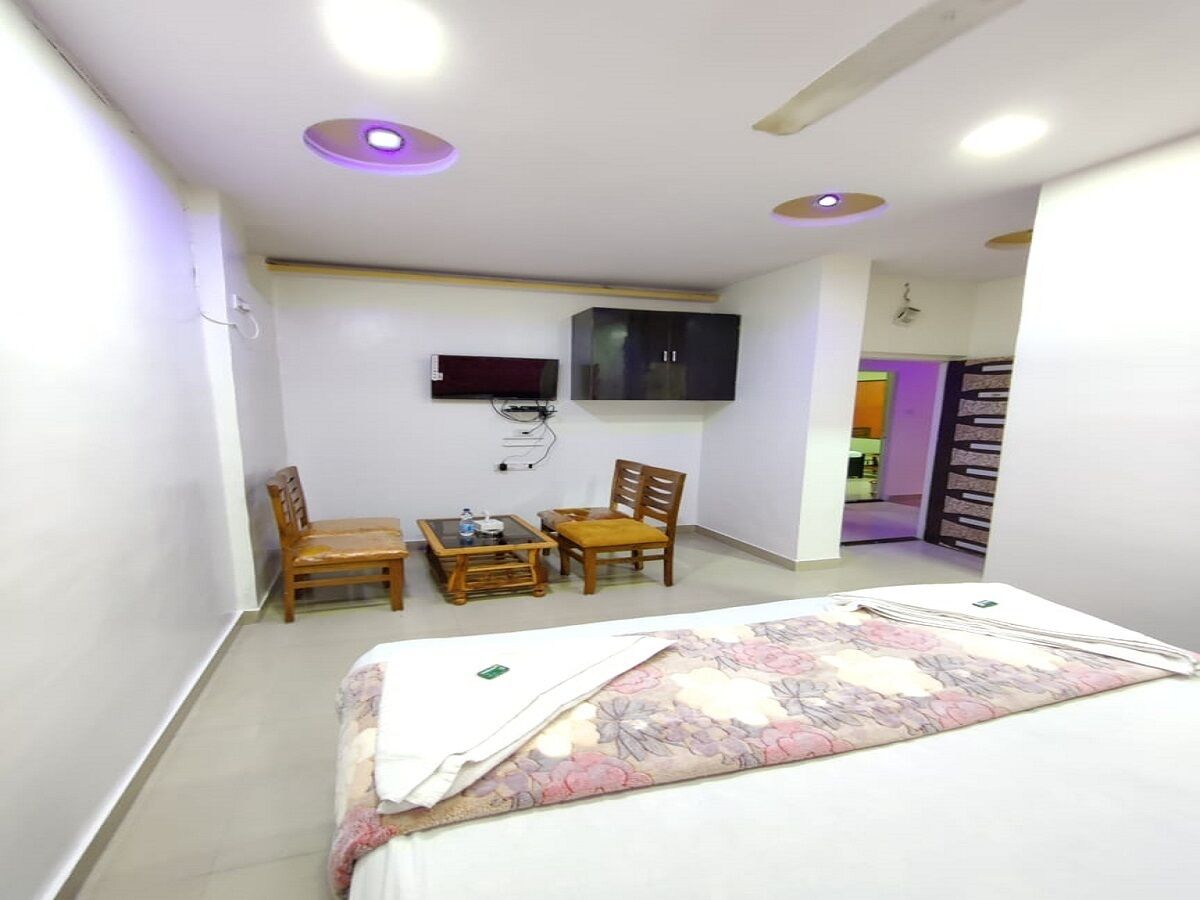 A bedroom with a bed, two wooden chairs, a small table, a TV mounted on the wall, and a door leading to another room.