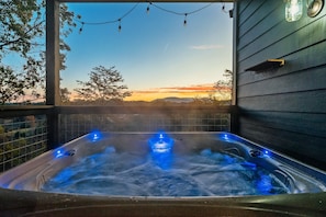 A hot tub with blue lighting, surrounded by a wooden deck and a view of a sunset. -  Brand New! • Views! • Easy Roads! • Hot Tub! • Beautifully Decorated! (Sevierville)