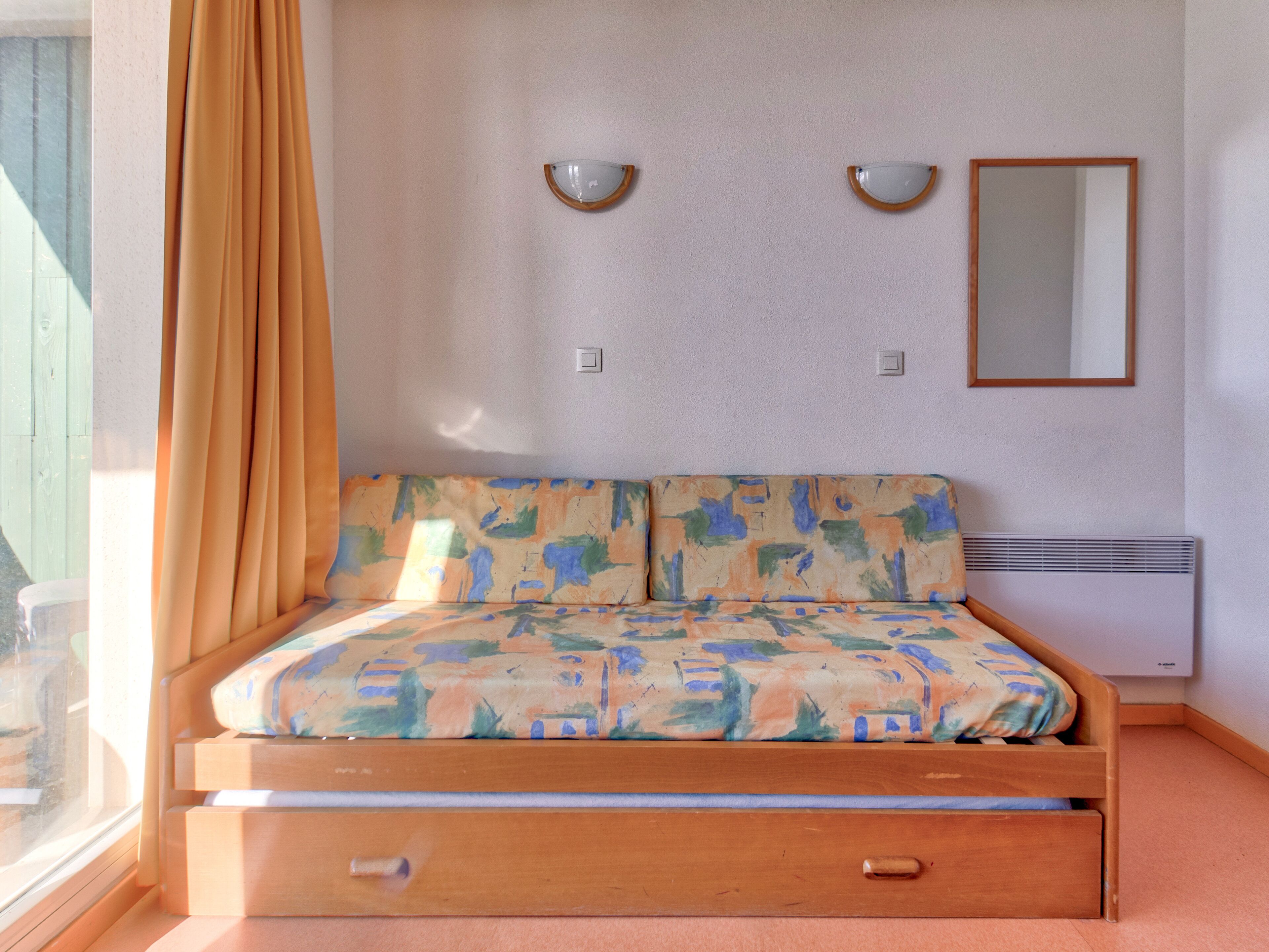 Een slaapkamer met een bed, een raam met gordijnen, een spiegel aan de muur en een wandheater.