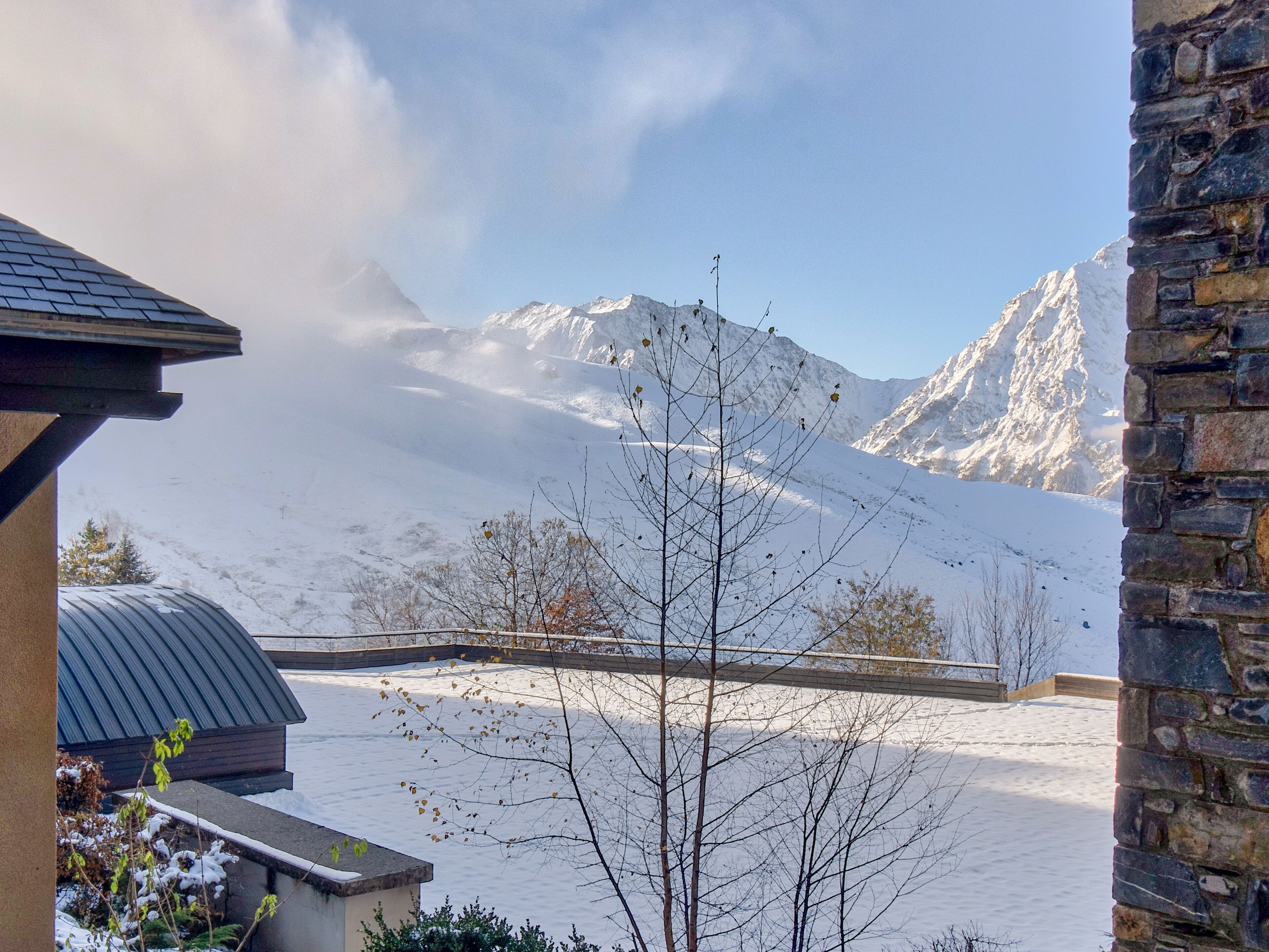 A snow-covered mountain landscape with a stone building and a metal roof structure in the foreground.