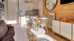 Una sala de estar moderna y bien iluminada, con un sofá gris, una pequeña mesa redonda con un jarrón de flores y una zona de cocina con armarios blancos y una pared de madera.