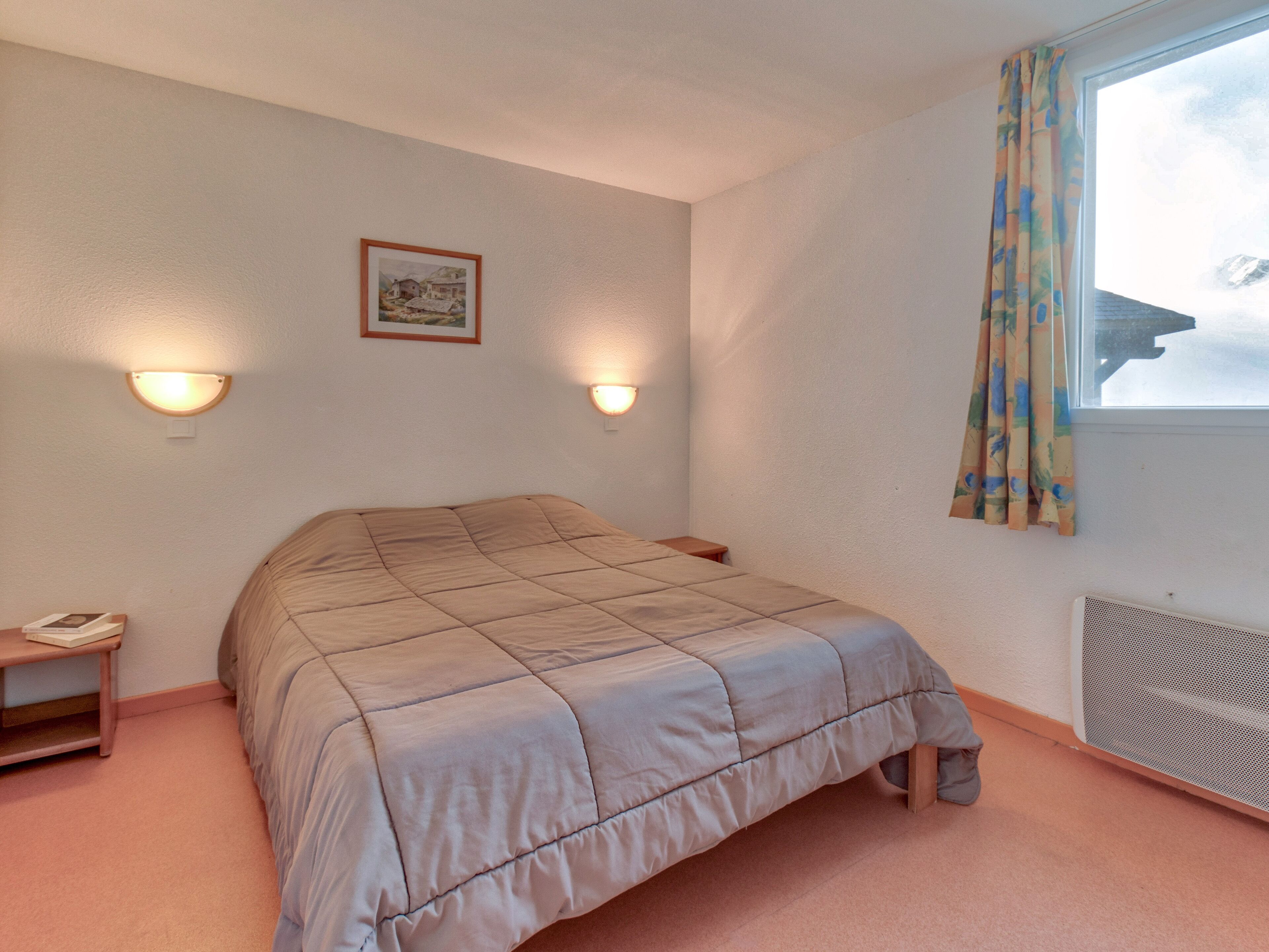Un dormitorio con cama, dos luces fijadas a la pared, una ventana con cortina y un radiador.