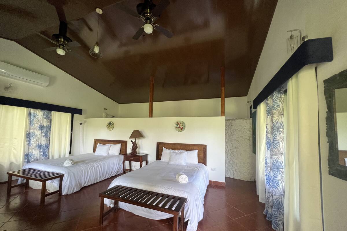 A hotel room with two beds, a ceiling fan, a small table, and a window with curtains.