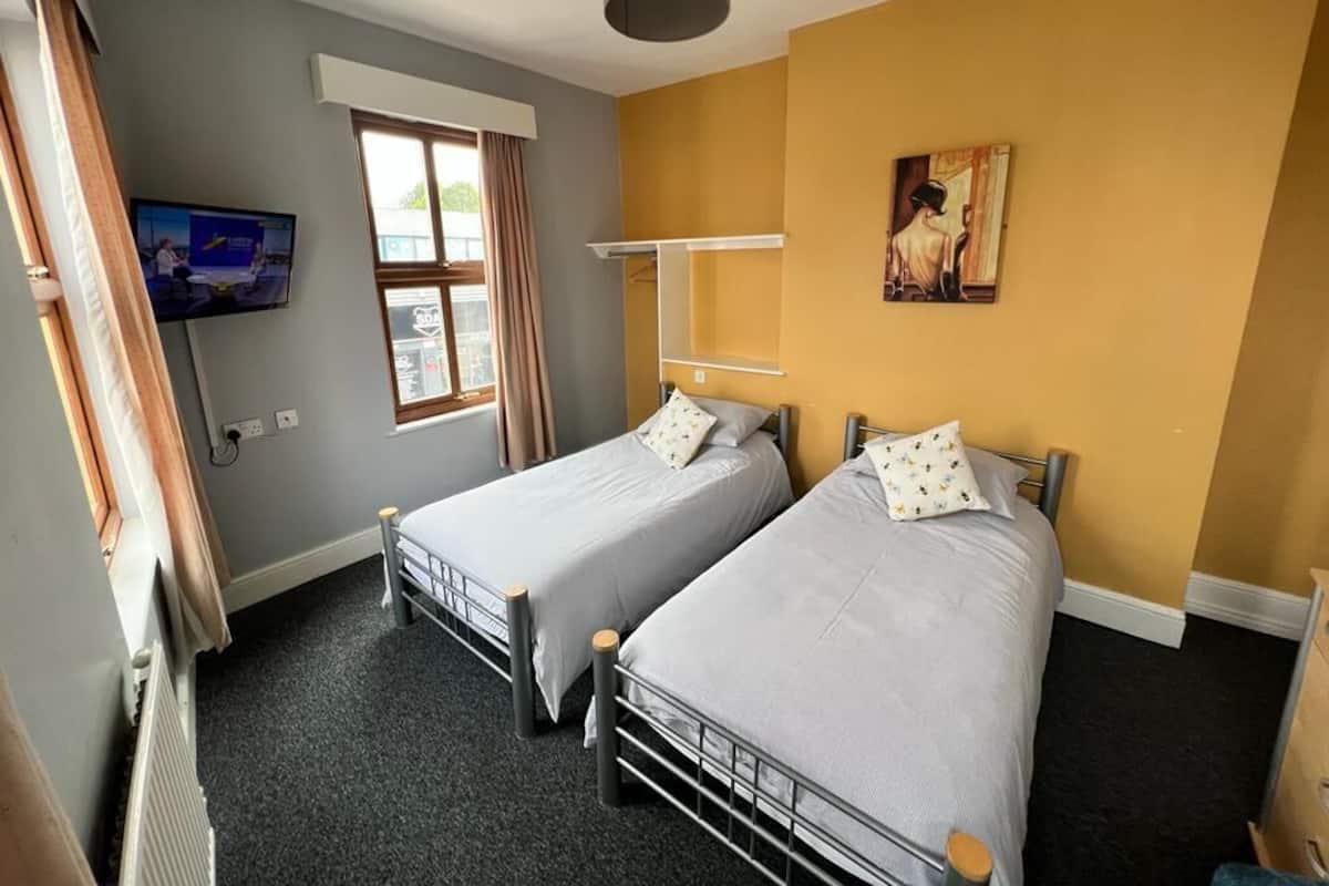 A room with two single beds, a TV mounted on the wall, a window with curtains, and a painting on the wall.